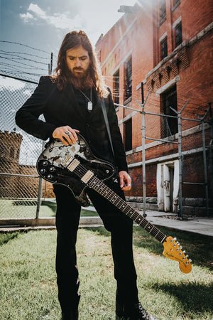 Tim Montana standing in the sun on a grassy, urban landscape. His hand is on his well-worn guitar, he is dressed in all black, and he is looking down at the ground.